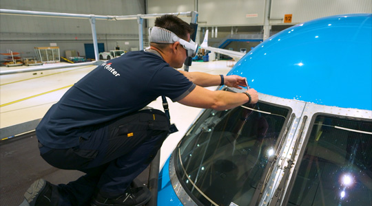 A technician in a plane hangar wears Apple Vision Pro while using a tool in their hands to adjust the plane’s windshield