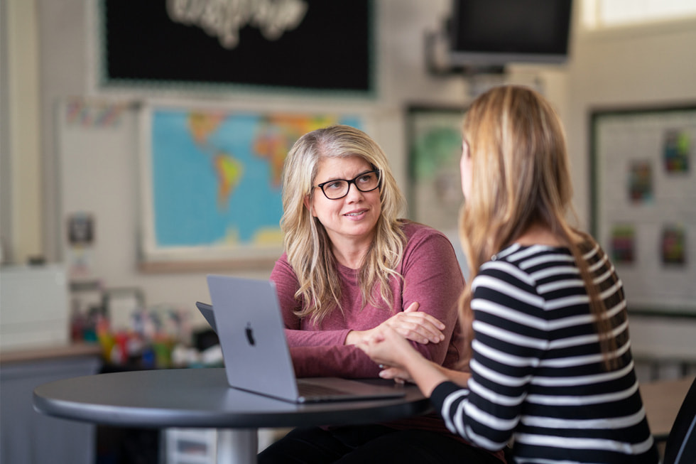 Downey Unified instructional technology coach Marnie Luevano speaks with another adult who is not facing the camera in a classroom setting, with MacBook Air on the table.
