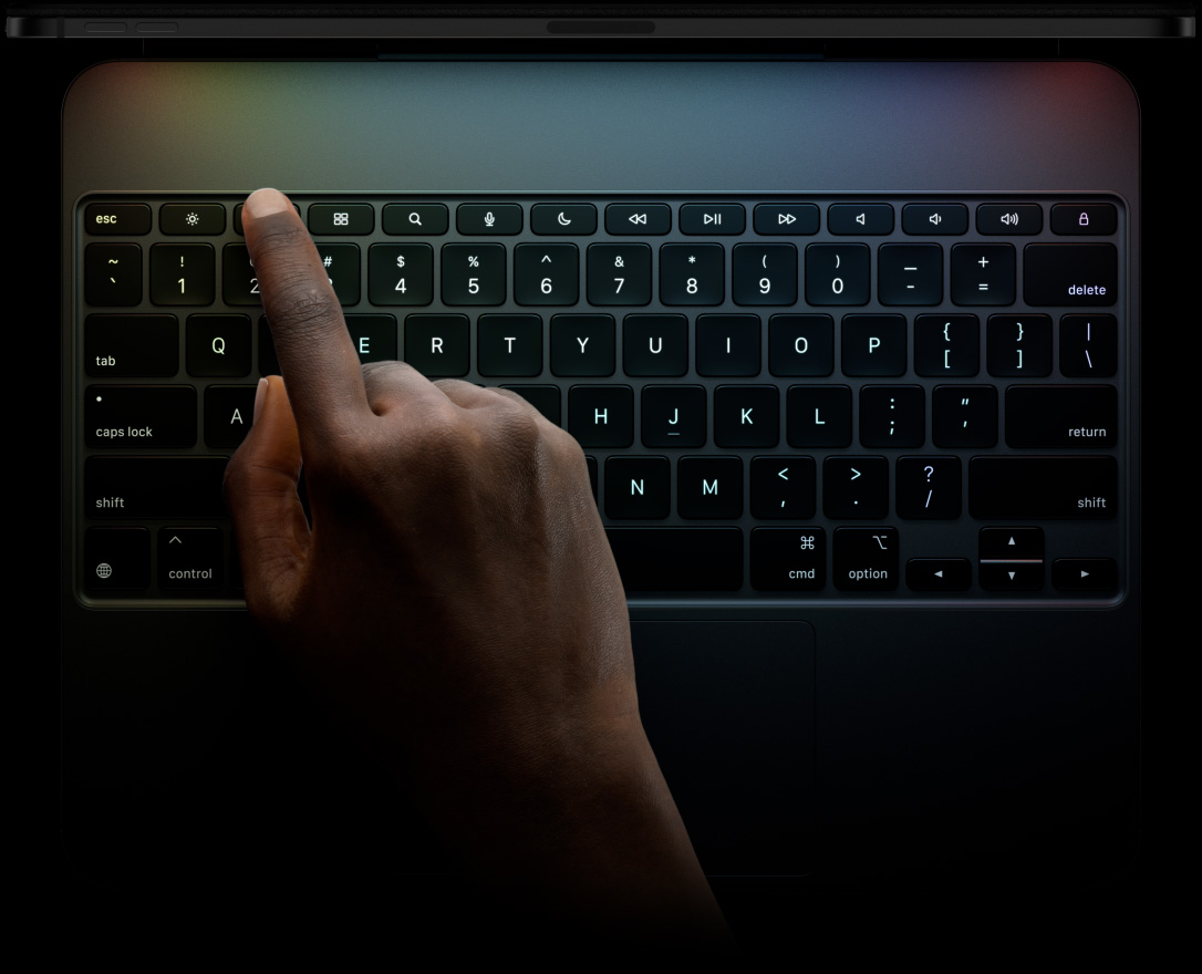 橫向放置的太空黑色 iPad Pro 頂部邊緣,貼合在橫向放置的巧控鍵盤上,鍵盤正面展示黑色背光按鍵、專用功能鍵列、escape 鍵、tab 鍵、大寫鎖定鍵、shift 鍵、control 鍵、option 鍵、shift 鍵、刪除鍵、return 鍵、以倒 T 形排列的方向鍵,以及內建觸控式軌跡板